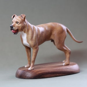 Puede incluir: Escultura de madera detallada de un perro marrón y blanco, con una pelota roja en la boca, sobre una base de madera oscura. La escultura está finamente elaborada, mostrando la musculatura del perro y sus rasgos realistas.