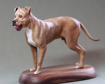 Puede incluir: Escultura de madera detallada de un perro marrón y blanco, con una pelota roja en la boca, sobre una base de madera oscura. La escultura está finamente elaborada, mostrando la musculatura del perro y sus rasgos realistas.