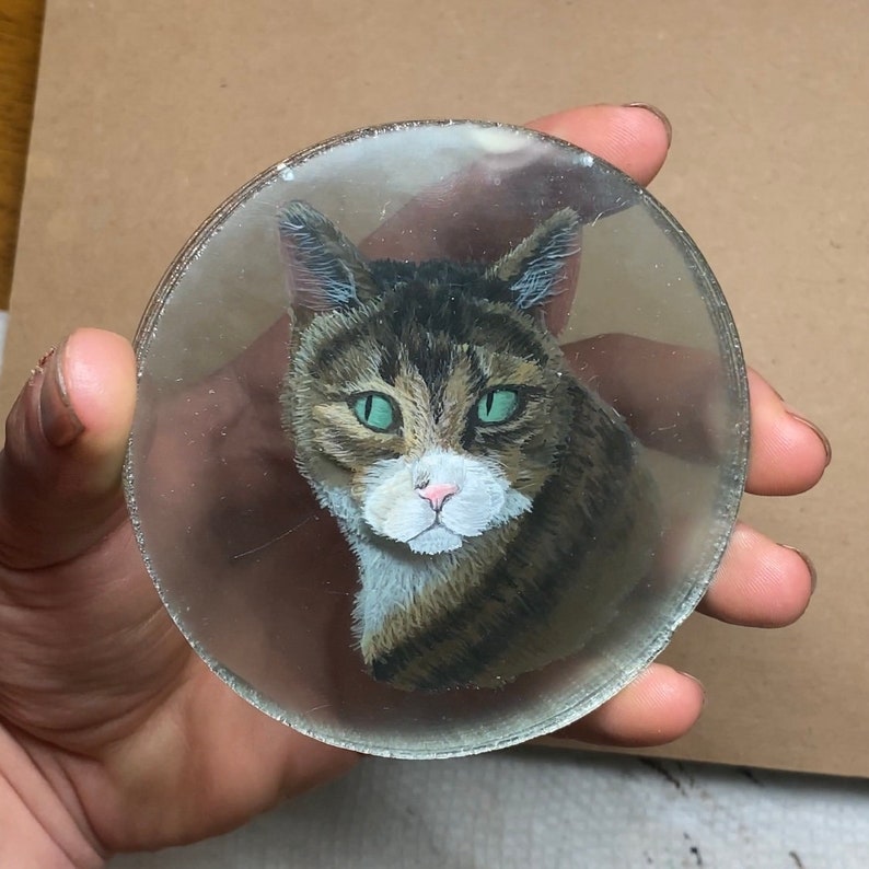 Puede incluir: Un primer plano de un retrato pintado de un gato atigrado con ojos verdes en un disco de vidrio transparente.