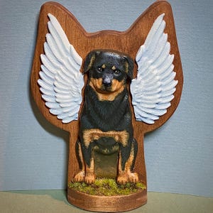 Puede incluir: Una talla de madera de un perro Rottweiler con alas de ángel blancas. El perro está sentado sobre una base de madera con musgo verde.