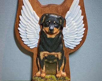 Puede incluir: Una talla de madera de un perro Rottweiler con alas de ángel blancas. El perro está sentado sobre una base de madera con musgo verde.