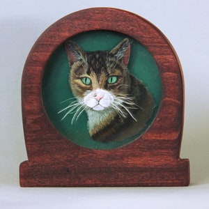 Puede incluir: Una pintura enmarcada de un gato atigrado con ojos verdes. El gato est&aacute; mirando directamente al espectador. El marco es de madera oscura y tiene una parte superior redondeada.