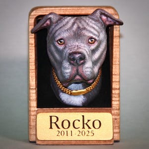 Puede incluir: Placa conmemorativa de madera con la cabeza esculpida de un perro gris y blanco con collar dorado. El nombre del perro, "Rocko", y los años "2011-2025" están grabados en una placa dorada en la parte inferior del marco.