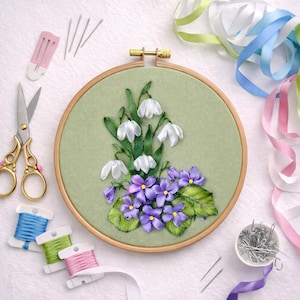 Può includere: Ricamo floreale con bucaneve bianchi e viole viola su tessuto verde chiaro, all'interno di un telaio di legno. Forniture per cucito, tra cui forbici, aghi, rocchetti di filo colorato e nastri, circondano il telaio.