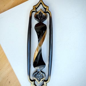 May include: A decorative door pull handle in a vintage style. The handle has a black and gold twisted central design, framed by a dark bronze-coloured border with floral accents at the top and bottom. The handle is designed for a door.