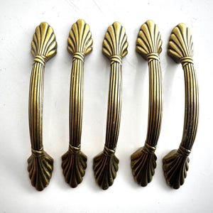 May include: Five antique brass cabinet pulls with a shell design. Each pull features a curved handle with vertical lines and decorative shell-shaped ends. The pulls are arranged on a white surface.