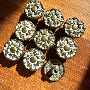 May include: A set of decorative cabinet knobs with a floral design. The knobs have a brass finish with cream-colored accents. They are arranged on a wooden surface, with one knob showing the mounting hardware. Each knob is approximately 4 cm in diameter.