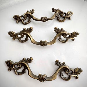 Peut inclure: Trois poignées de tiroir antiques en laiton avec des motifs floraux ornés et défilants. Les poignées ont une poignée incurvée et des extrémités décoratives, mettant en valeur une esthétique vintage. La finition en laiton a une patine chaude et vieillie, ajoutant à leur attrait classique.