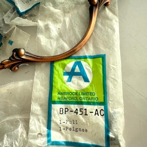 May include: A copper-colored cabinet pull with an ornate, curved design. The pull is packaged in a clear plastic bag with a label that reads "AMEROCK LIMITED MEAFORD, ONTARIO" and "BP-451-AC".
