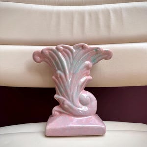 May include: A ceramic vase featuring a glossy finish in shades of pastel pink and soft green. The object has a wide, flared top with curved, leaf-like ridges and a solid, rectangular base. The surface has a smooth, reflective, glazed texture.