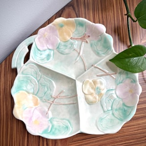 May include: A divided serving dish in the shape of a leaf, with a handle on the side. The dish is light green with pink and yellow floral accents. The dish is made of ceramic.