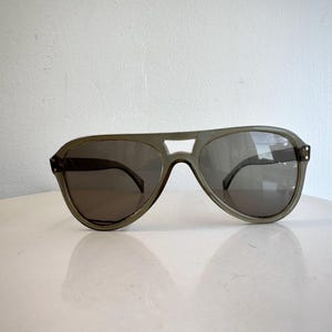 May include: Aviator-style sunglasses with a gray frame and dark lenses. The sunglasses have a distinctive brow bar and a classic teardrop shape. The lenses are dark, providing shade and protection from the sun. A stylish accessory for any outfit.