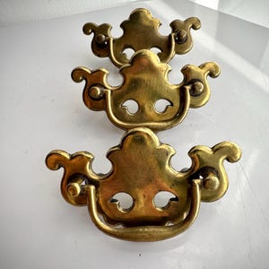 May include: Three antique brass drawer pulls with a decorative, ornate design. Each pull features a curved handle and a stylized, symmetrical shape with two eye-shaped cutouts. The brass has a warm, aged patina.