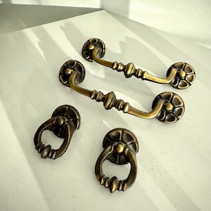 May include: Four antique brass drawer pulls and handles. Two are long, with a curved handle and decorative floral ends. The other two are smaller, ring-shaped pulls with similar detailing. All have a warm, aged finish.