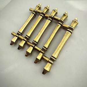 May include: Six vintage brass cabinet pulls. Each pull features a curved central handle with decorative ends and a crossbar at each end. The brass has a warm, aged patina. The pulls are arranged in two rows of three.
