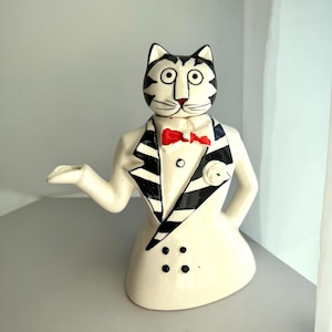May include: A ceramic cat figurine wearing a tuxedo with a red bow tie. The cat has black and white stripes on its head and a white body. The tuxedo has black lapels and three black buttons. A small white flower is pinned on the lapel.