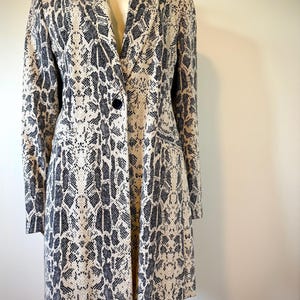 May include: A long, beige and black snakeskin-patterned blazer. The blazer features a single black button closure, long sleeves, and two front pockets. The garment is displayed on a mannequin.
