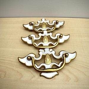 May include: Three white and gold drawer pulls with a decorative design. The pulls have a curved handle and ornate detailing. The hardware is set against a light wood surface.
