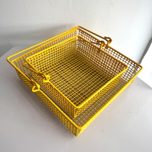 May include: Two bright yellow metal wire baskets, one slightly smaller than the other, are nested together. Each basket has a rectangular shape with a grid pattern and a handle on each side. The baskets are on a light grey surface.