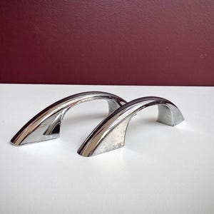 May include: Two arched cabinet pulls with a polished chrome finish. Each handle features a smooth, curved design with tapered ends that slope down to the mounting points, creating a sleek, reflective metallic appearance for furniture hardware.