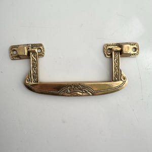 May include: A brass drawer pull with decorative scrollwork and a curved handle. The pull has two mounting plates with screw holes and a detailed design on the handle. The brass finish gives it a vintage look.