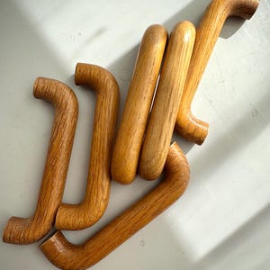 May include: Six wooden furniture handles in a light brown hue, arranged on a white surface. Each handle features a curved design and visible wood grain. The handles are likely for cabinets or drawers.