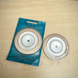 May include: Two round white ceramic knobs with gold-tone metallic rims and a central square mounting hole. One knob is attached to a blue cardboard backing card with the text "CERAMOID, WHITE & GOLDEN ACCENT" printed at the bottom.
