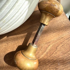 Peut inclure: Bouton de porte ancien en laiton avec un dessus et un dessous arrondis, reliés par une tige métallique rectangulaire. Le laiton présente une patine chaude et vieillie, suggérant un charme vintage. Le bouton est probablement pour une porte ou une armoire.