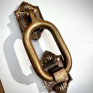 Peut inclure: Un heurtoir de porte en laiton antique avec une poignée ovale et une plaque arrière ornée. La plaque arrière présente un motif décoratif à volutes. Le heurtoir est de couleur laiton vieilli et chaleureux, suggérant un charme vintage.