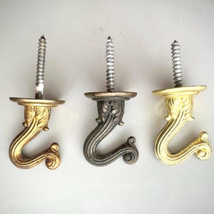 May include: Three ornate metal screw-in hooks. The hooks have a decorative, curved design with a floral motif. One hook is gold, one is silver, and one is a darker, antique-looking gold. Each hook has a silver screw.