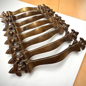 May include: Eight antique bronze drawer pulls are displayed. Each pull features a curved handle and ornate end details. The bronze has a warm, aged patina. The pulls are arranged on a white surface.
