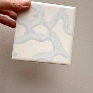 May include: A square ceramic tile with a glossy white finish featuring a light blue abstract pattern of interconnected, organic, branch-like shapes that extend across the surface. The tile has smooth, straight edges and a flat, reflective face.