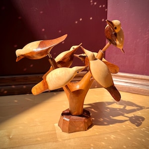 May include: A decorative wooden sculpture featuring five carved birds perched on a central branch structure. The birds are crafted from two-toned light and dark wood, mounted on a faceted, dark brown wooden base with a smooth, polished finish.