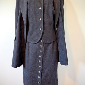 May include: A dark grey pinstripe skirt suit. The suit comprises a blazer, a button-down vest, and a long skirt. The blazer has a notched collar and the skirt has buttons down the front. The suit is displayed on a mannequin.