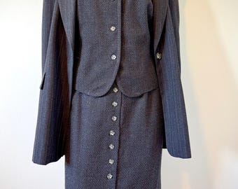 May include: A dark gray pinstripe skirt suit. The suit includes a blazer, a button-down vest, and a long skirt. The blazer has a notched collar and the skirt has buttons down the front. The suit is displayed on a mannequin.