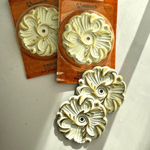 Peut inclure: Médaillons décoratifs floraux ivoire et or. Les médaillons présentent un motif orné avec un trou central. Deux sont emballés sur des cartes marron avec le logo "Amerock" et des informations sur le produit.