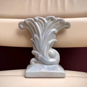 May include: A light blue ceramic vase with a glossy finish, featuring a flared, fan-like top and a sculpted, swirling base. The object has a smooth, reflective surface and a wide, rectangular bottom that provides a stable foundation for the piece.
