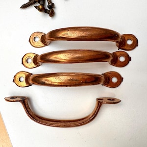 May include: Four antique copper-coloured drawer pulls and several screws are arranged on a white surface. Three pulls are arched with rounded ends and two screw holes. The fourth pull is curved with two mounting plates.