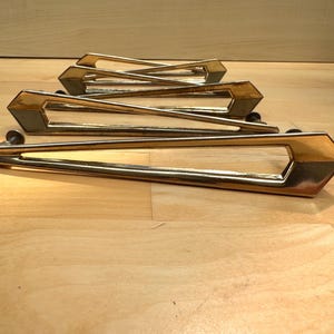 May include: A set of three elongated, gold-toned metal drawer pulls. Each handle features a sharp, angular, geometric design with a hollow centre and two mounting points on the underside, arranged in a row from the foreground to the back.
