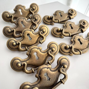 May include: A collection of antique brass drawer pulls. Each pull features a keyhole and decorative scrollwork. The hardware has a warm, aged finish, suggesting vintage or retro styling. The pulls are arranged on a white surface.