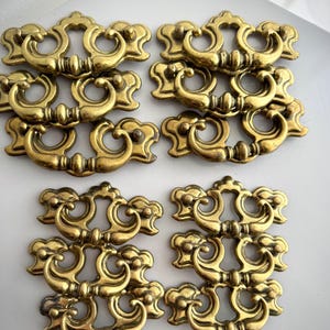 May include: Eight ornate brass drawer pulls. Each pull features a decorative design with a curved handle and intricate detailing. The pulls are arranged in two rows of four, showcasing their vintage style and potential for furniture restoration projects.