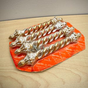 May include: Four cabinet pulls in a twisted rope design with a white and gold finish. The hardware is sealed in original orange plastic packaging featuring the text Charm n Style, Banner Industries, 181 Carlaw, Toronto, and M4M 2S1.