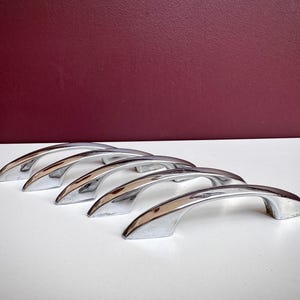 May include: A set of five polished chrome cabinet handles arranged in a row. Each handle features a smooth, arched design with tapered ends and a reflective metallic finish. The hardware is constructed with a curved grip and two mounting points.