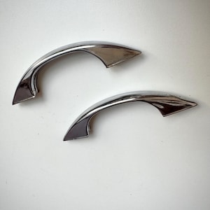 May include: Two matching silver-toned metal drawer pulls with a curved, arched design. Each handle features a smooth, reflective finish and tapered ends that flare out slightly at the mounting points, creating a sleek and functional hardware piece.
