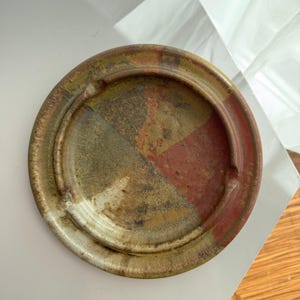 May include: A circular ceramic ashtray with a segmented design. The ashtray has a raised rim and is divided into four colour sections: red, green, and yellow-brown. The ashtray has a rustic, handmade appearance.