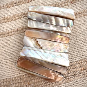 May include: A bracelet made of rectangular, iridescent shell pieces. The colours vary from pearly white to warm brown, with hints of pink and green. The bracelet is displayed on a textured, beige surface.