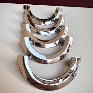 May include: Five polished chrome cabinet pulls arranged in a vertical stack. Each pull features a semi-circular, crescent-shaped design with a smooth, reflective metallic finish. The hardware is uniform in size and shape, showcasing a curved arc.