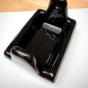 May include: A glossy black ceramic soap dish featuring a rectangular basin with rounded corners and a raised back section containing a single circular hole for wall mounting. The interior is smooth and deep, designed to hold a bar of soap securely.