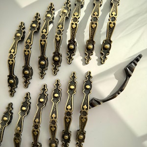 May include: A collection of antique-style cabinet handles. The handles are dark bronze with gold accents, featuring an ornate, symmetrical design. The handles are arranged on a white surface, showcasing their decorative details.