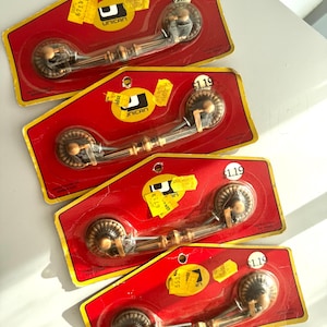 May include: Four vintage bronze-toned drawer pulls on red and yellow packaging. Each pull has a decorative circular end and a curved handle. The packaging includes the text "UNICAN" and price tags.